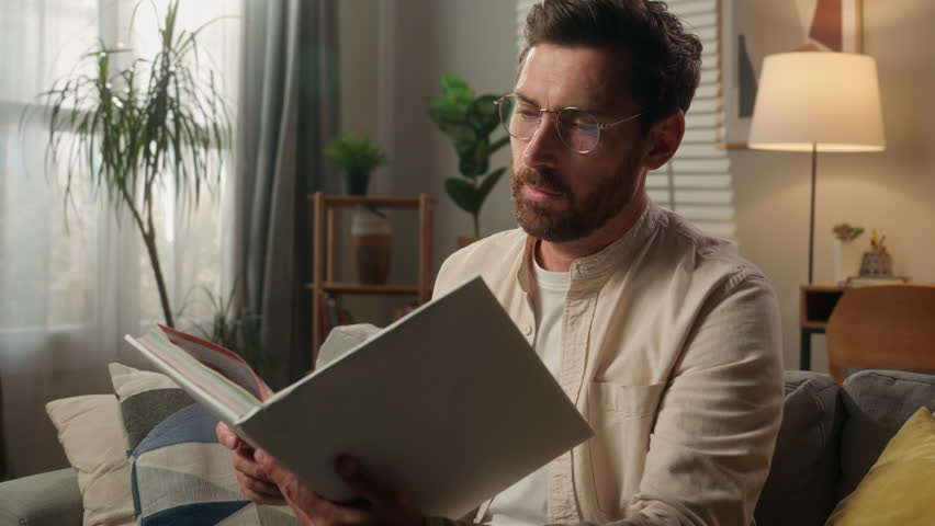 Caucasian man intelligent male guy on couch sofa reading book alone at home adult homeowner reader businessman in glasses read literature paper textbook hobby leisure in apartment studying knowledge