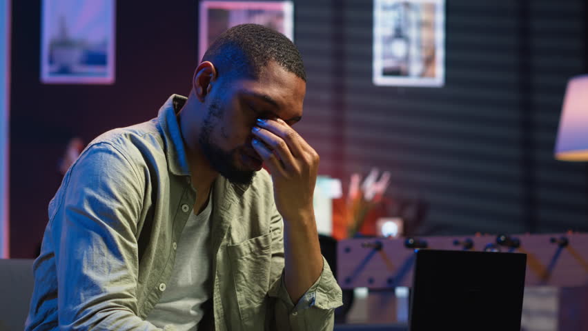 African american person receiving some bad news while doing remote work tasks, feeling overworked and under pressure to complete all his assignments. Young adult deals with burnout. Camera B.