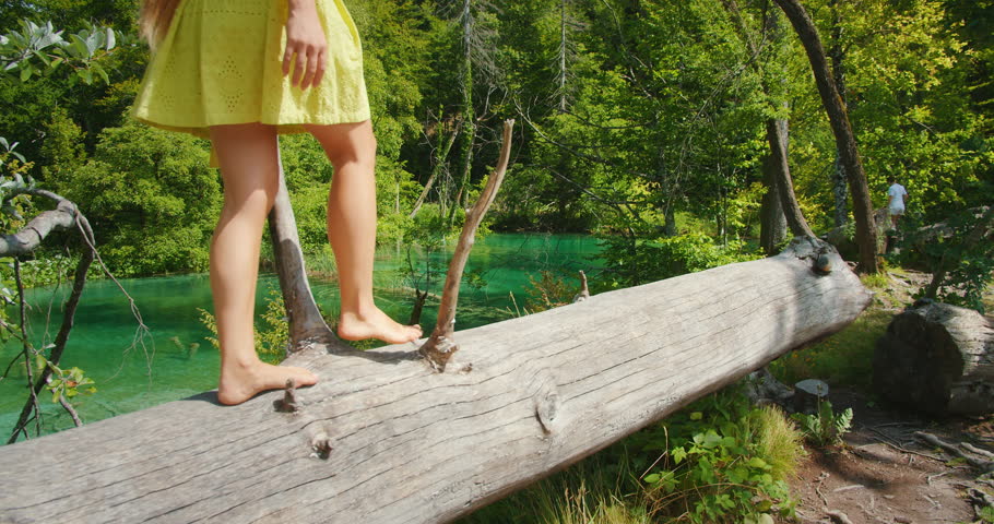 Barefoot woman in dress balances on fallen log by Plitvice turquoise lake in Croatia