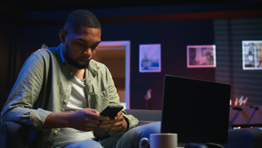 Relaxed black man scrolling on his smartphone in a comfortable space, sitting at the coffee table. Modern home highlights leisure and online connectivity in a stylish apartment. Camera A.