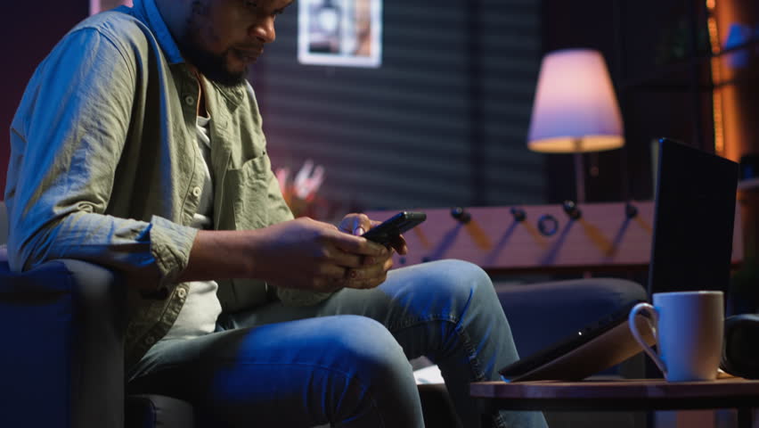 Relaxed black man scrolling on his smartphone in a comfortable space, sitting at the coffee table. Modern home highlights leisure and online connectivity in a stylish apartment. Camera B.