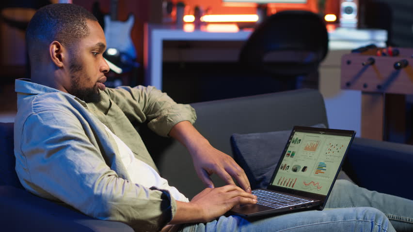 Black male trader monitoring stock market real time changes on laptop, investing funds with the goal to boost earnings. Focused young guy studying economic values for global advancement. Camera B.