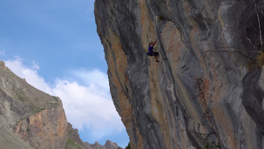 LOW ANGLE VIEW: Female rock climber scales an overhanging limestone cliff in an alpine environment. Fearless woman is secured with rope and wearing climbing gear as she climbs up a dramatic rock face.