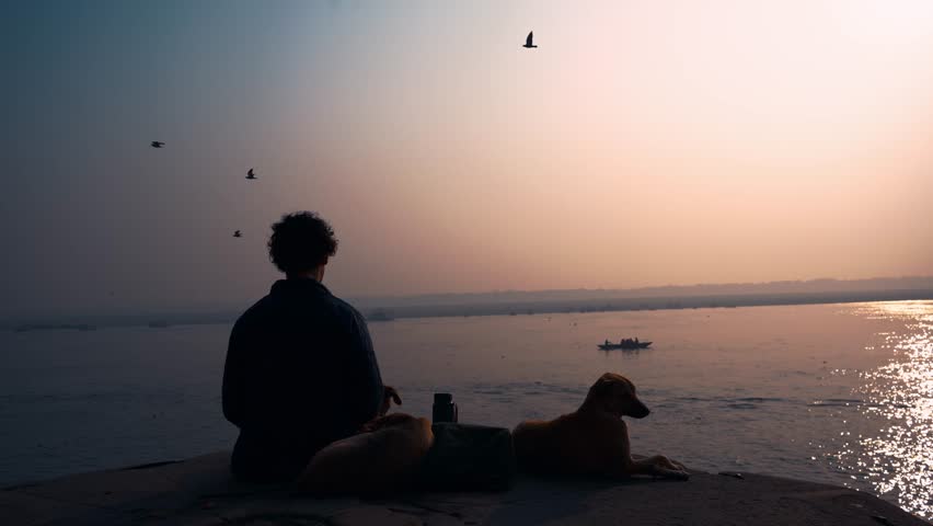 Man meditating at sunrise on a ghat by the sacred Ganges River, with dogs resting nearby. Peaceful spiritual moment in India with warm morning light and tranquil atmosphere.
