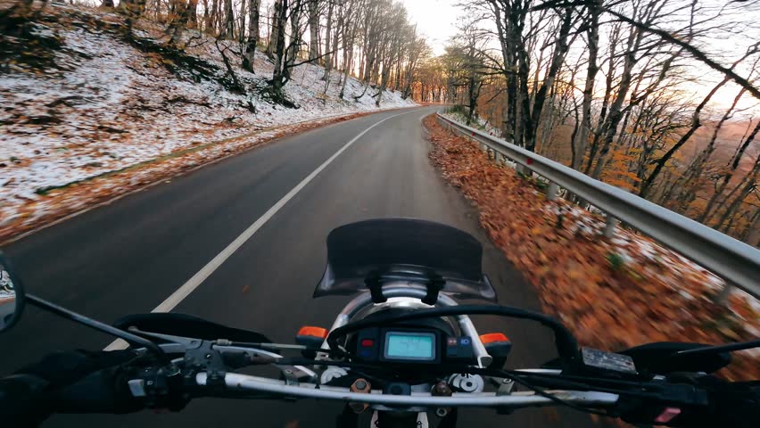 POV motorcycle ride through a scenic autumn forest on a paved road. Early frost, snow on the sides, golden sunset light shining through the trees.