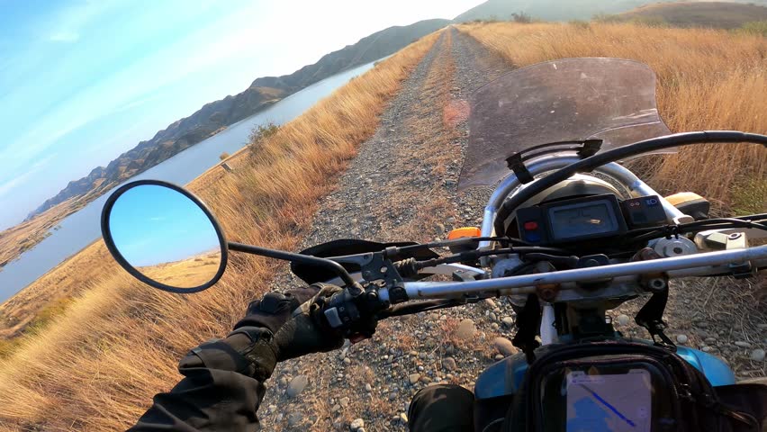 POV motorcycle ride through golden mountain fields at sunset, riding along a scenic alpine lake. Warm sunlight, peaceful nature, adventure and freedom on two wheels