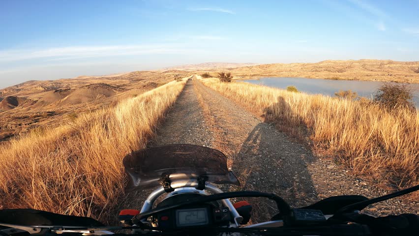 POV motorcycle ride through golden mountain fields at sunset, riding along a scenic alpine lake. Warm sunlight, peaceful nature, adventure and freedom on two wheels