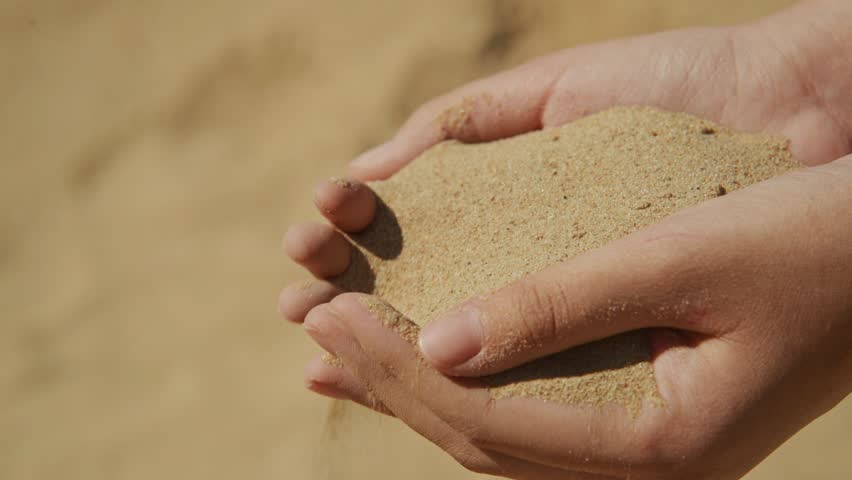 Two hands pour warm beach sand, creating a gentle cascade. Sunlight shines on the golden grains, capturing the feeling of relaxation and enjoyment by the shore.