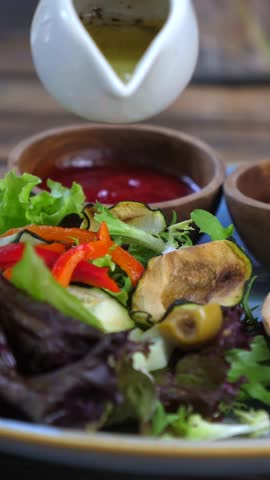 Vertical screen olive oil and spices cascading over fresh mixed salad with grilled vegetables, served alongside ketchup in wooden bowls