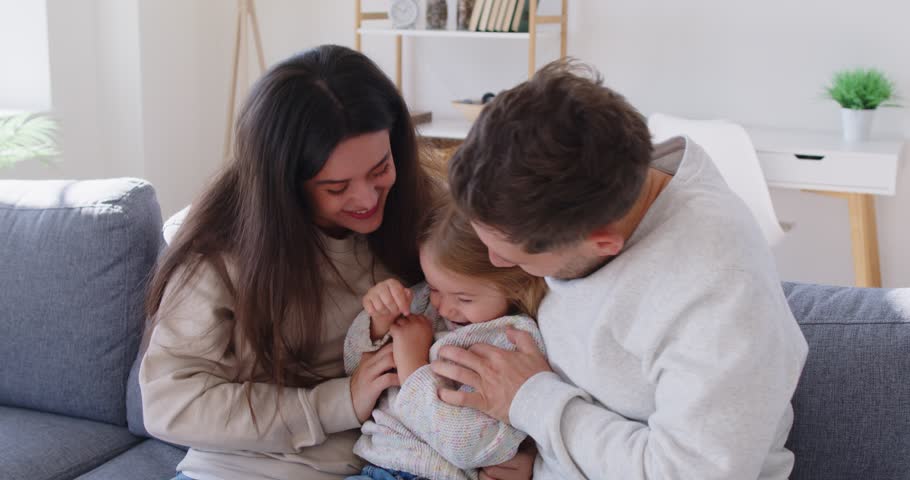 Mother and father play with their daughter at home tickling and having fun with her. Parents of little girl create precious family memories while sitting on sofa at home. Happy family concept.