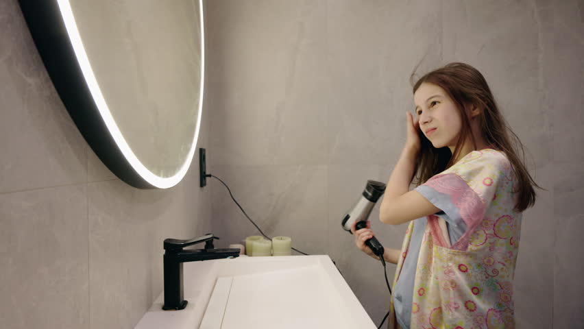 Teenage girl carefully drying her hair with a hairdryer in front of a mirror during her morning beauty routine at home. High quality 4k footage