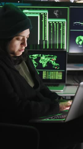 Vertical HD video of professional female hacker typing virus code on laptop for cyber attack in dark room with multiple monitors on the background
