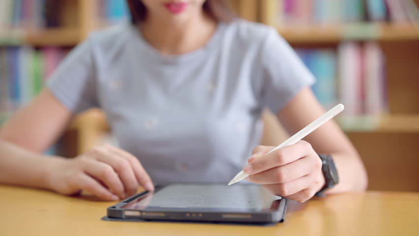 A woman is writing on a tablet with a pen. She is sitting at a table with a stack of books behind her - Powered by Shutterstock - Get 15% off with code: PIKWIZARD15