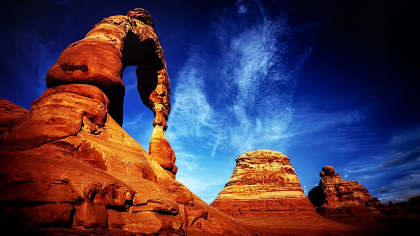 Sunset light over Delicate Arch in Utah, USA. Striking time lapse with high clouds and pure blue sky. Towering rock formations, orange layers of sandstone enhancing the majestic atmosphere.