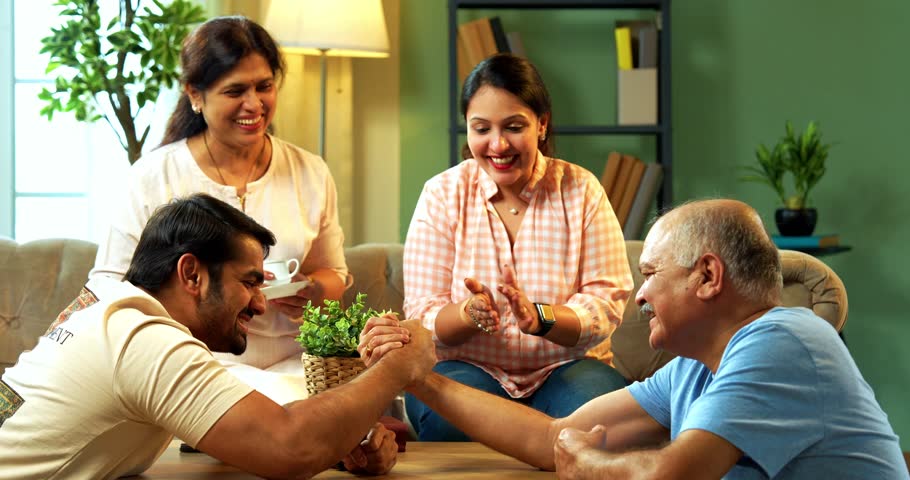 Arm wrestling fun between Indian senior father and young adult son in cozy living room while senior wife and young daughter laugh and cheer, enjoying friendly playful family moment at home together