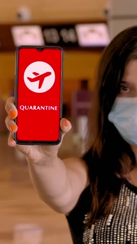 Woman, in protective mask, holds smartphone with airplane icon, text - quarantine, on screen, in front of empty check-in counters at the airport.