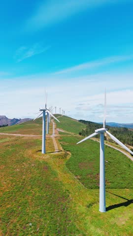 Aerial view of large white wind turbines with spinning blades on top of a mountain. Wind power generating self-sufficient, eco-friendly electricity on a beautiful sunny summer day.
