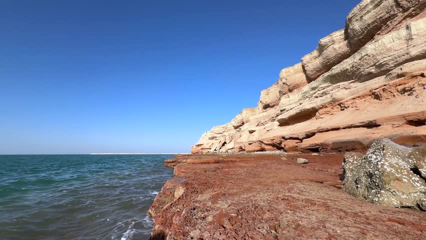 Turquoise waters washing crimson rocky shores of Hormuz Island, exposing unique geological formations beneath clear azure skies in Persian Gulf landscape