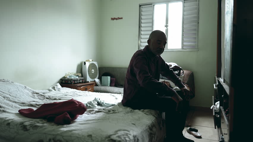 One pensive serious middle-aged man seated by bedside in a moody dark room struggling with poverty and depression, thoughtful person of African descent