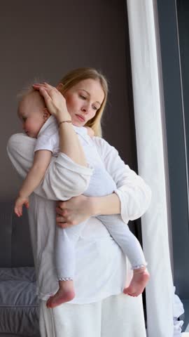 Beautiful mom gently embracing baby near window at home, in natural light, dressed in white casual clothes, enjoying tender family indoors on bright day. Mum with little child, daughter