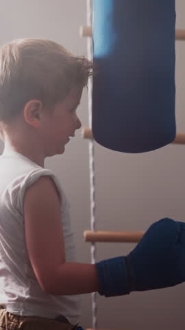 Curious boy in blue gloves learns to punch bag at professional boxing class. Sportive toddler enjoys boxing training in modern gym slow motion