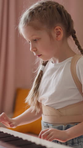 Elder sister with plaits shows piano chords to little brother and asks to repeat at home. Toddler boy learns to play music with schoolgirl in living room