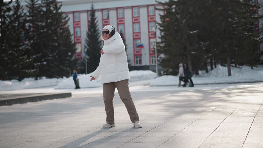 Elegant woman in white coat performing kung fu-inspired moves on snowy park pavement while people walk in background near statue and tall pine trees in cold urban setting with clear winter sky