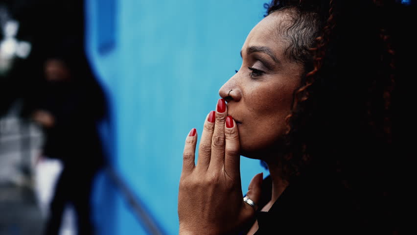 One pensive worried middle-aged black latina hispanic woman in deep contemplative dilemma standing in urban street leaning on blue wall struggling with depression
