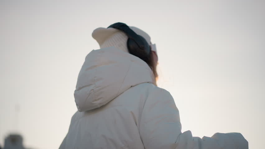 Shoulder view of creative woman smiling as she glides through park in winter outfit, wearing headphones and beanie, enjoying moment under clear sky with warm light casting gentle glow