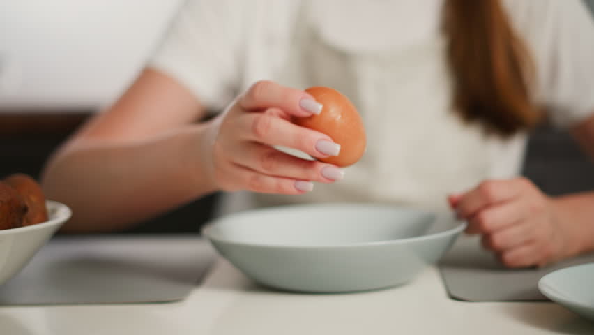 Close up of woman cracking egg shell on glass plate then peeling shell from boiled egg over bowl at kitchen table, wearing apron with potatoes and dishes in background, showing cooking prep