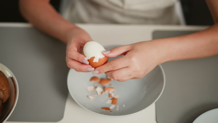Overhead view of fair skinned person focused on peeling shell off boiled egg over bowl on table with scattered shell fragments, on placemat beside potatoes and empty dish