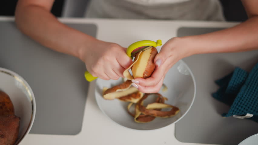 Aerial view of young woman hands peeling sweet potato over white bowl filled with potato skins, placed on dining table with towel and side bowl, capturing moment of focused food preparation