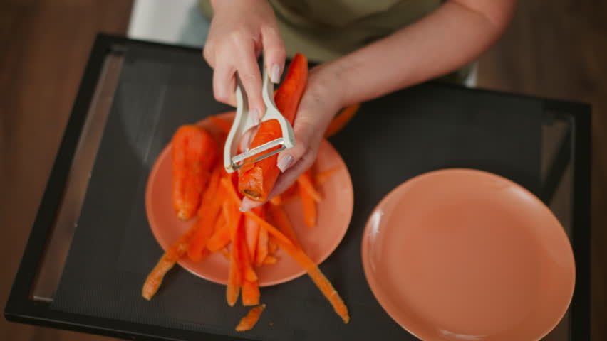Overhead view chef in green shirt peeling fresh carrot with white peeler over black tray, bright orange peels piled on plate beside empty plate on wooden floor under soft natural light