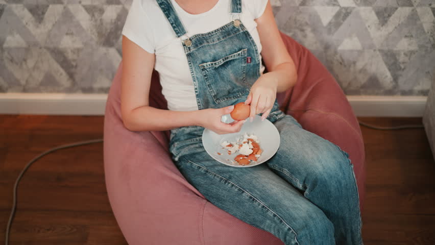 Overhead view of person in blue denim overall seated on pink bean bag carefully peeling egg shell into white bowl placed on lap, with relaxed posture and scattered egg shells