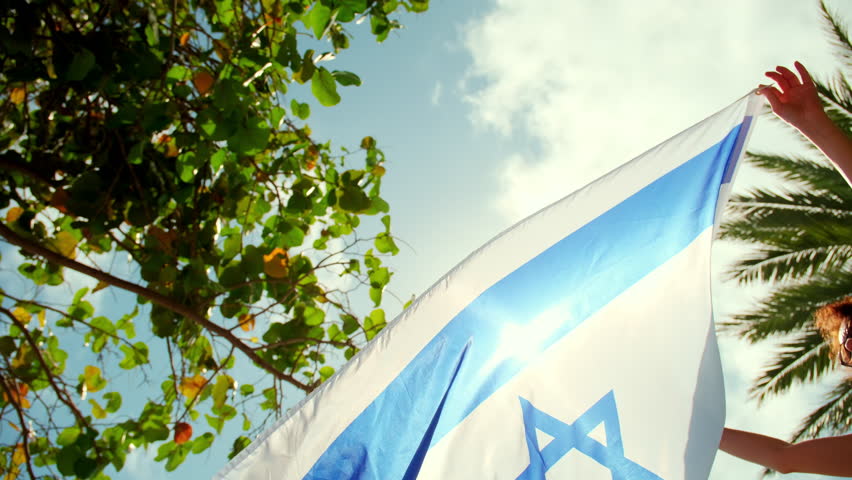 Woman holds the Israel flag against a strong wind, with a background of clear blue sky and tropical trees.