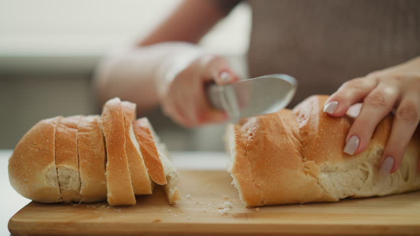 Close up of female chef cutting loaf of bread with knife on wooden board, slicing into equal parts under natural light, focus on hands guiding blade across crust and forming uniform slices