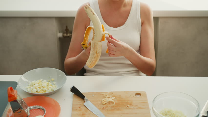 White lady removing banana peel carefully in bright kitchen during food preparation, sitting at table with cutting board, knife, grated ingredients, and other kitchen tools visible around workspace
