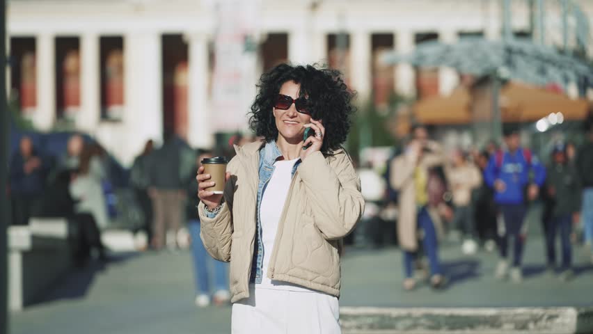 Bussinesswoman make mobile communication walking in city and smiling, chat or biracial female entrepreneur in conversation, discussion or negotiation holding a cup of coffee on sunny day, slow motion
