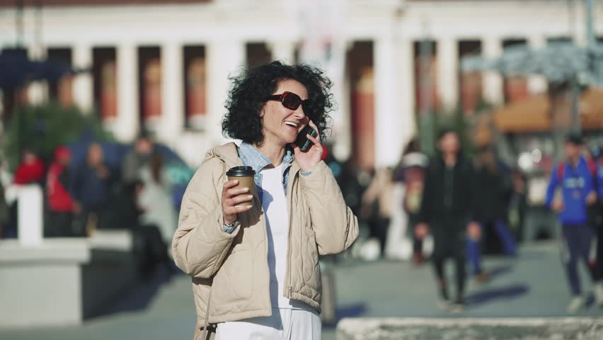 Bussinesswoman make mobile communication walking in city and smiling, chat or biracial female entrepreneur in conversation, discussion or negotiation holding a cup of coffee on sunny day, slow motion