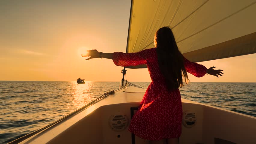 Girl in red dress travel on ocean. Young woman is sailing on yacht under sail her hands raised dream of fly over ocean. Lady travel on ocean on yacht watching sunrise. Ship sailing sea woman looks sun