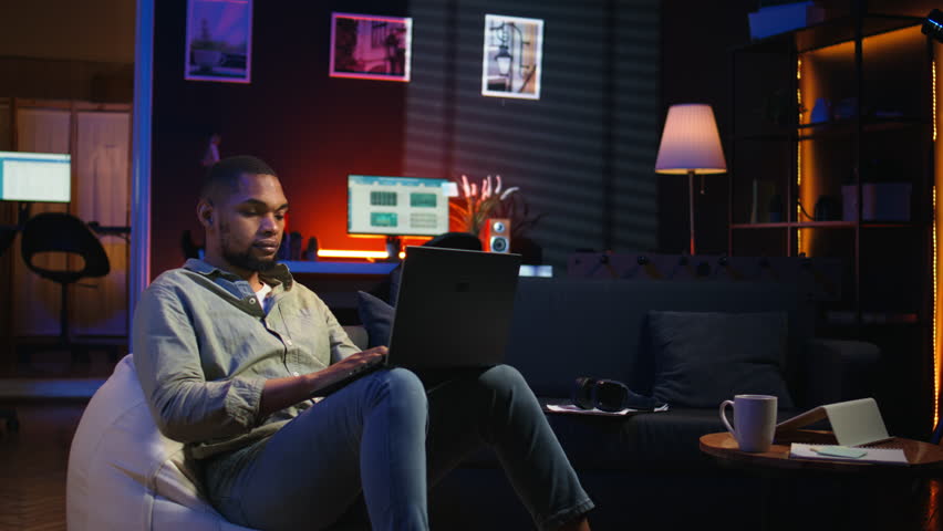 African american man working remotely from his modern loft on laptop, focusing on tasks completion while he lays on a bean bag. Blending productivity and relaxation, telework. Camera A.