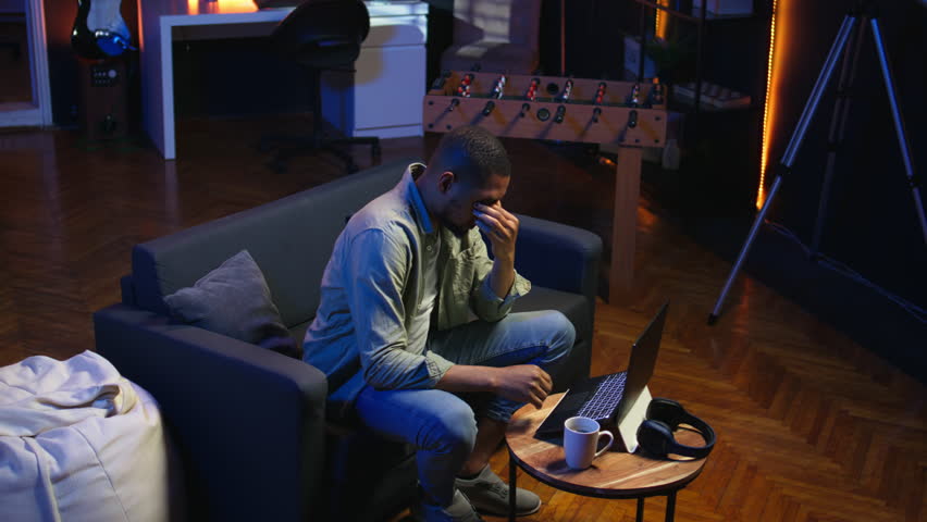 African american person receiving some bad news while doing remote work tasks, feeling overworked and under pressure to complete all his assignments. Young adult deals with burnout. Camera A.
