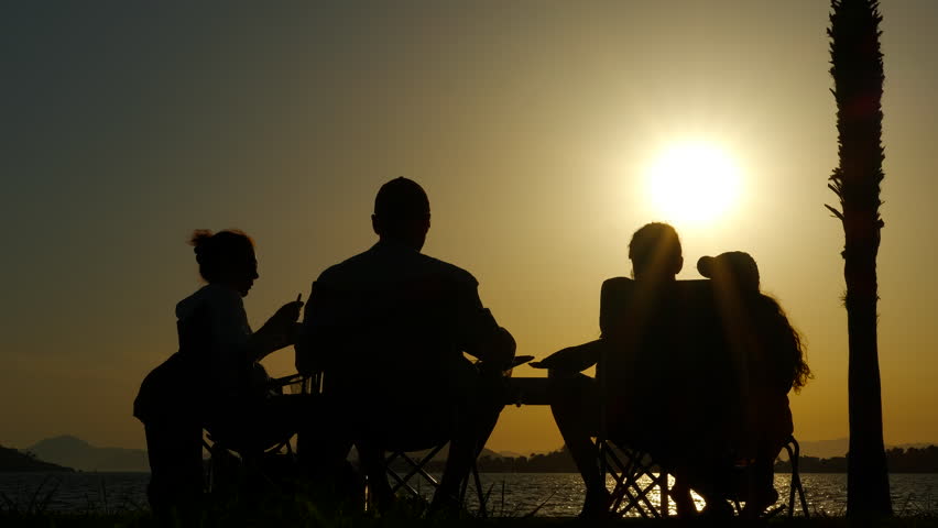 Silhouetted figures of a family enjoying a relaxing picnic by the lake create a heartwarming scene as the sun sets, casting a golden glow across the tranquil sky