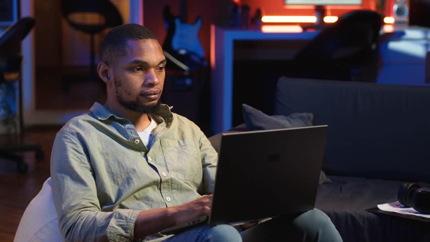 African american man working remotely from his modern loft on laptop, focusing on tasks completion while he lays on a bean bag. Blending productivity and relaxation, telework. Camera B.