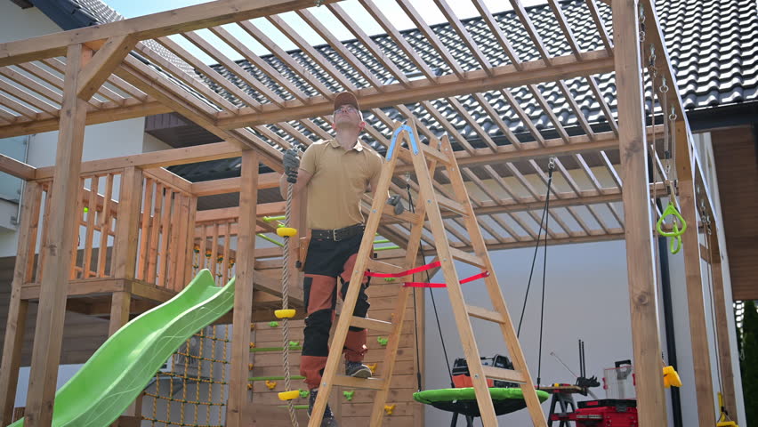 A skilled individual is actively assembling a wooden playset in a backyard setting. The sunlight shines down on the area, highlighting the detail of the structure. Set atop a sturdy ladder the builder