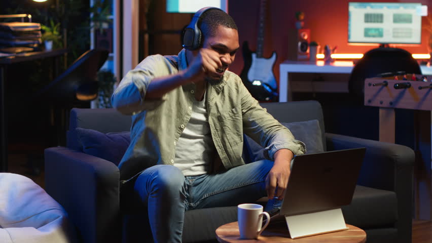Focused black guy teleworking on laptop in a living room feels happy with task completion, achieving success. Remote worker balancing comfort and work efficiency in a flexible workspace. Camera B.