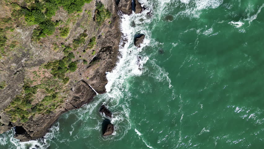 Top-down drone shot showing waves crashing onto rocky coastline near Piha, New Zealand. Green water and rugged shore.