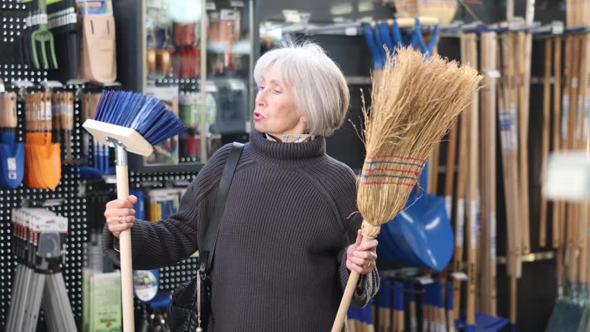 Mature woman with gray hair chooses a broom and a large brush to clean the garden. Senior woman at the sale of equipment in a garden store. High quality 4k footage