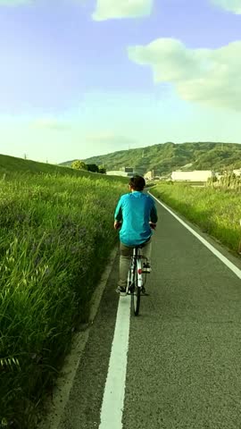 A young man cycles alone on a country road lined with green grass, embracing freedom, simplicity, and the joy of a quiet journey.
Japan, 03 May,2025.