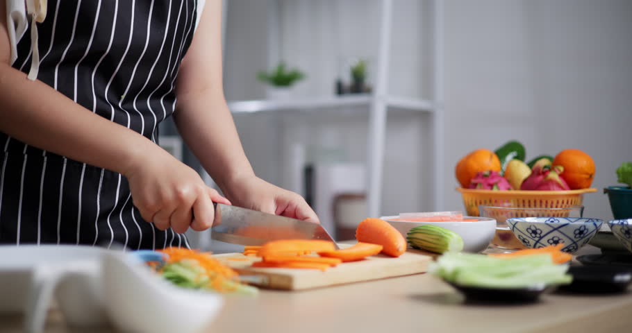 Handheld selective focus shot, Woman's hands cutting carrots with knife on wooden cutting board, Asian woman preparing fresh vegetables to make salad rolls. 